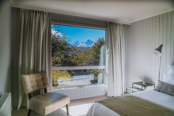 Un dormitorio con una ventana grande, una cama, una silla, una mesa y una lámpara.