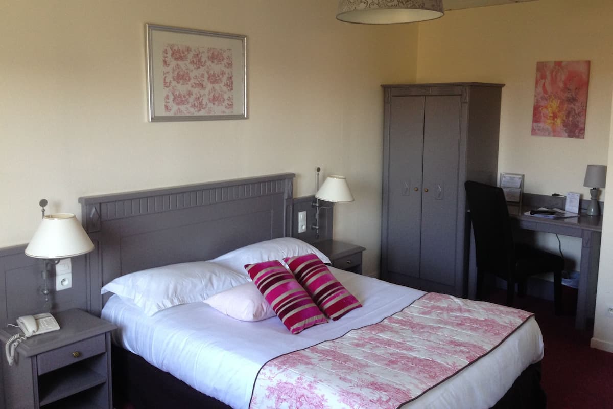 A hotel room with a bed, two bedside lamps, a desk with a chair, a wardrobe, and a hanging lamp.
