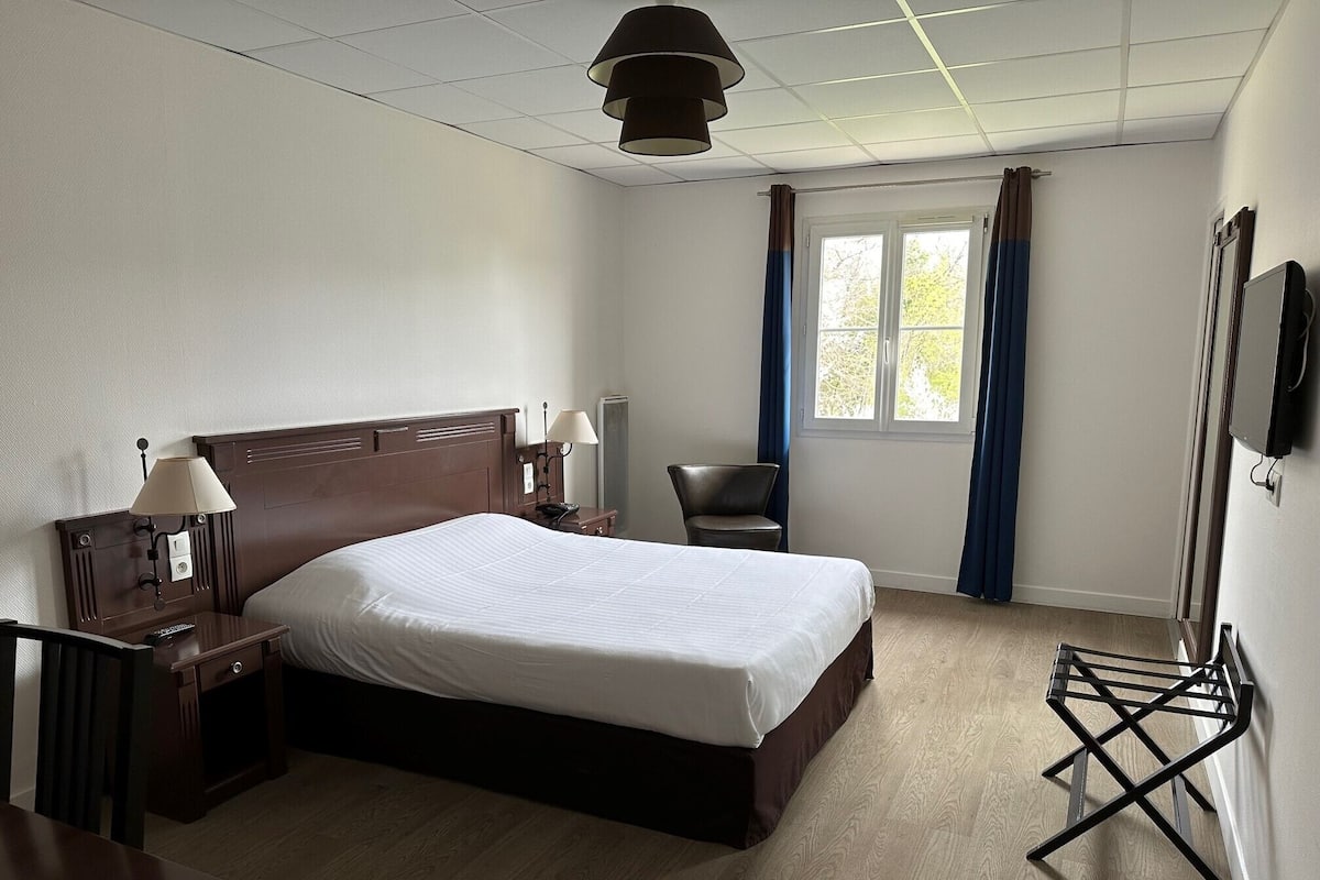 A hotel room with a large bed, bedside lamps, a desk, a chair, a TV, and a window with curtains.