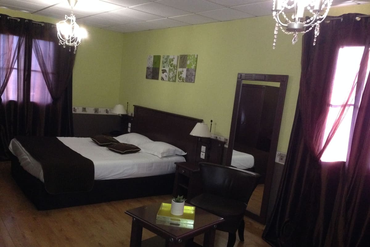 A hotel room with a large bed, a bedside table, a chair, and a small table. There are curtains, a chandelier, and artwork on the wall.