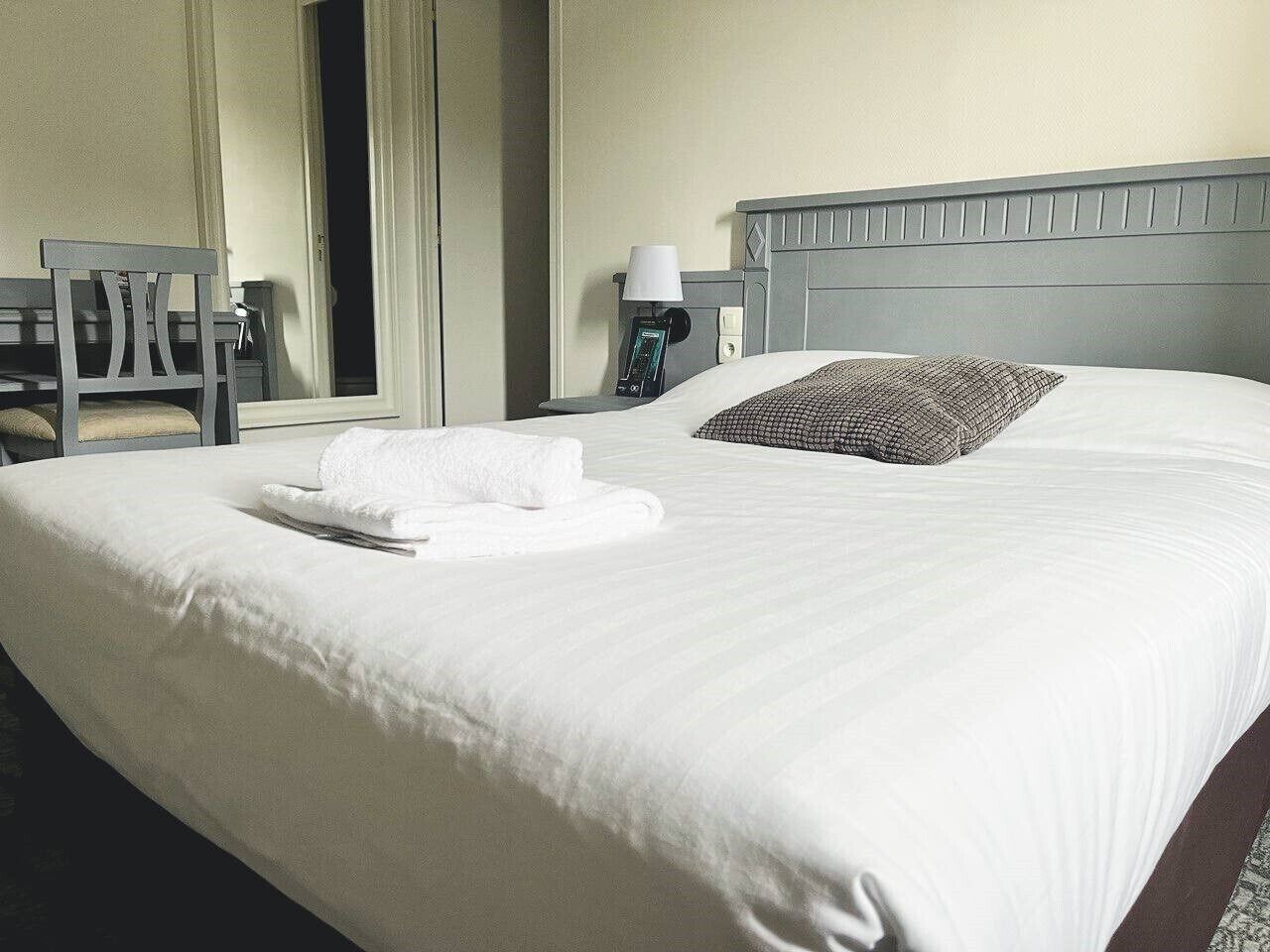 Una cama bien tendida con sábanas blancas y una toalla doblada, cabecera gris, lámpara de noche y una silla al fondo.