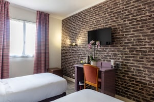 A hotel room with two beds, a desk, a chair, a TV mounted on a brick wall, and a window with curtains.