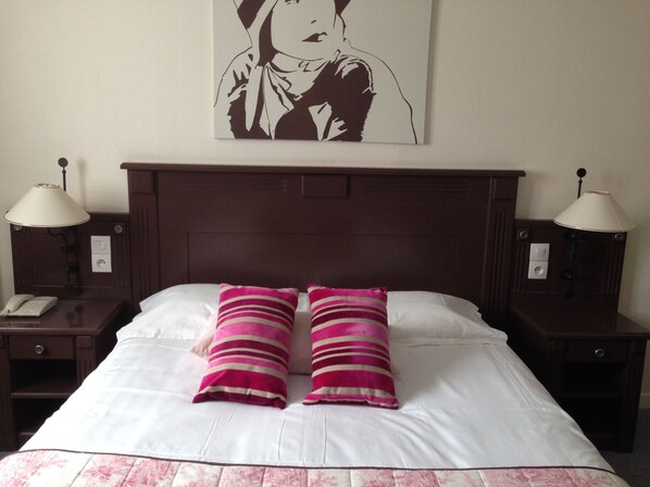 Habitación de hotel con una cama grande, dos lámparas de noche, una mesita de noche con un teléfono y una obra de arte colgada en la pared.