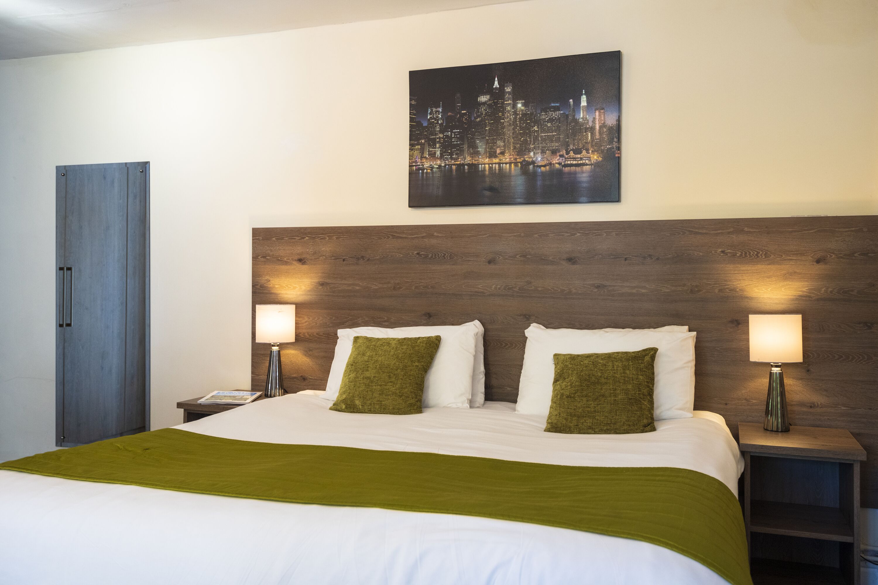 A hotel room with a large bed, two bedside lamps, a wooden headboard, a cityscape painting, and a wardrobe.