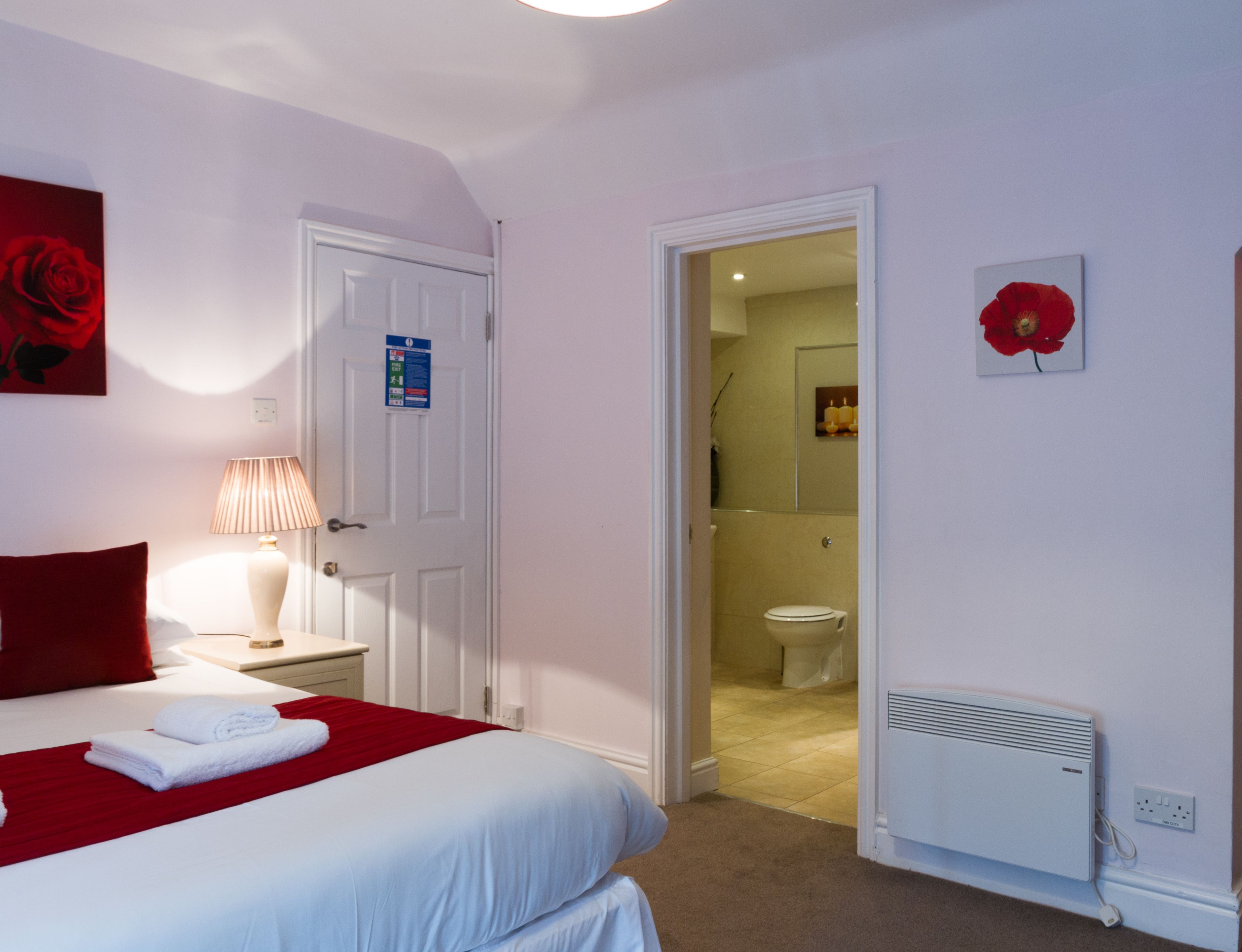 A hotel room with a bed, a nightstand with a lamp, a red flower decoration, and a white radiator.