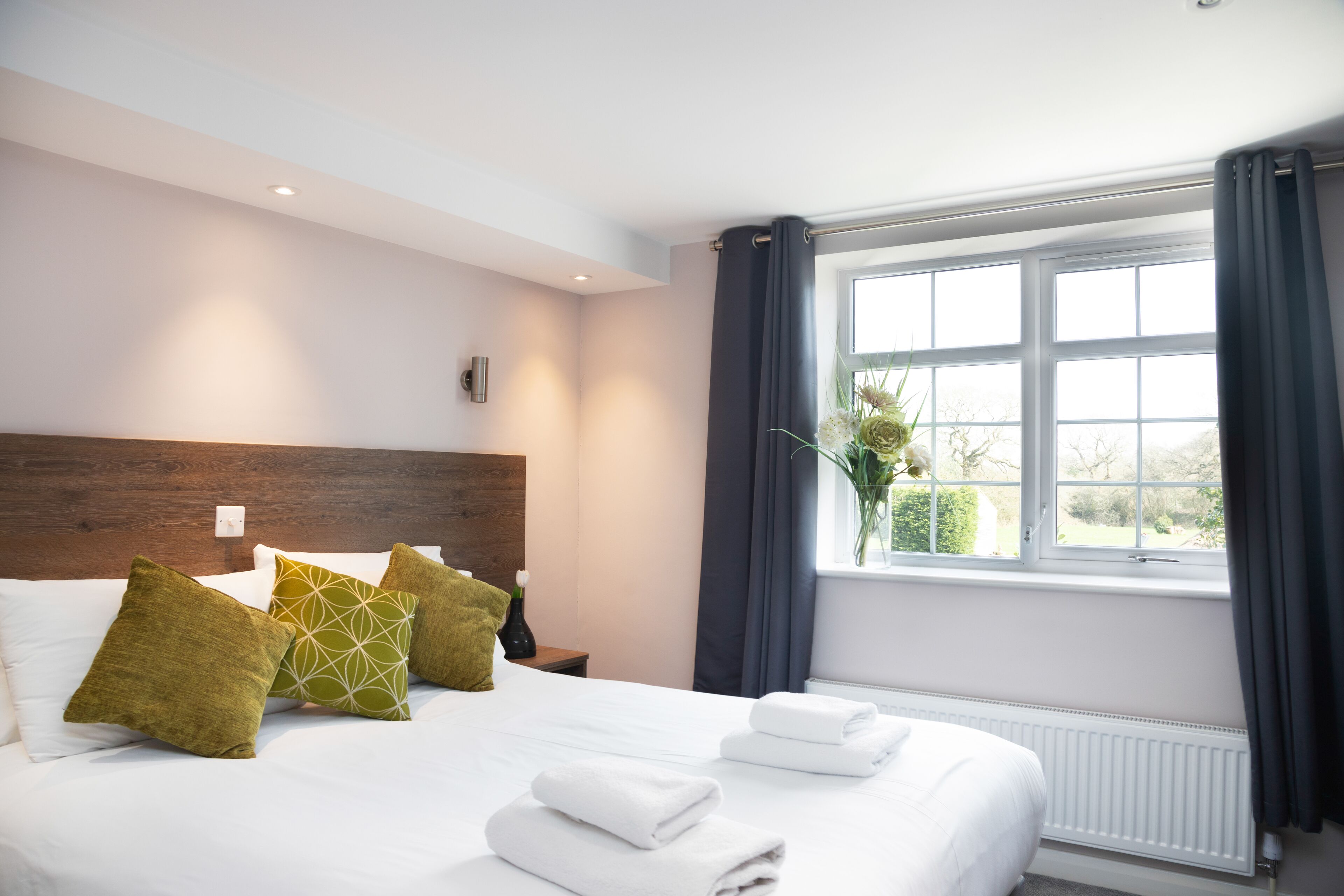 A neatly made bed with white linens and green patterned pillows, a wooden headboard, and a window with curtains.