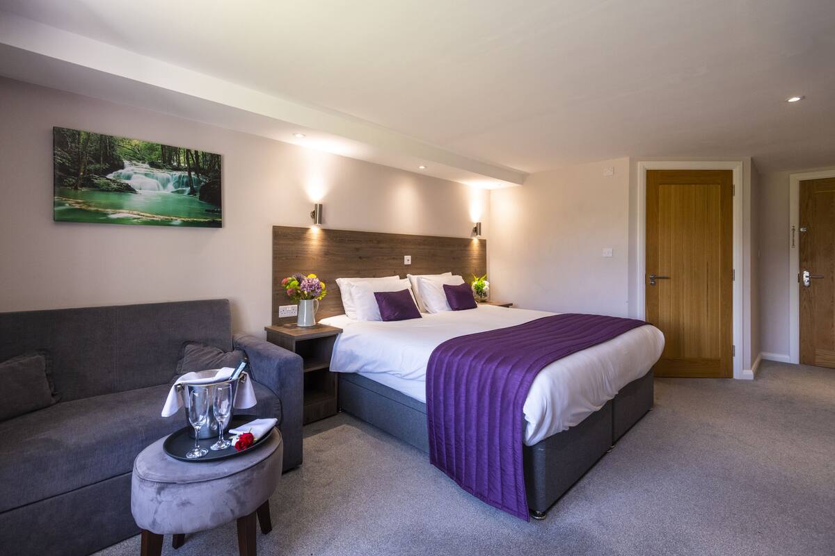 A modern hotel room with a large bed, a grey sofa, a small table with a tray, and a painting of a waterfall on the wall.