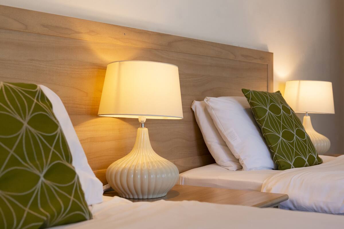 A hotel room with two beds, each with a white pillow and a green patterned pillow. There are two bedside lamps with white shades.
