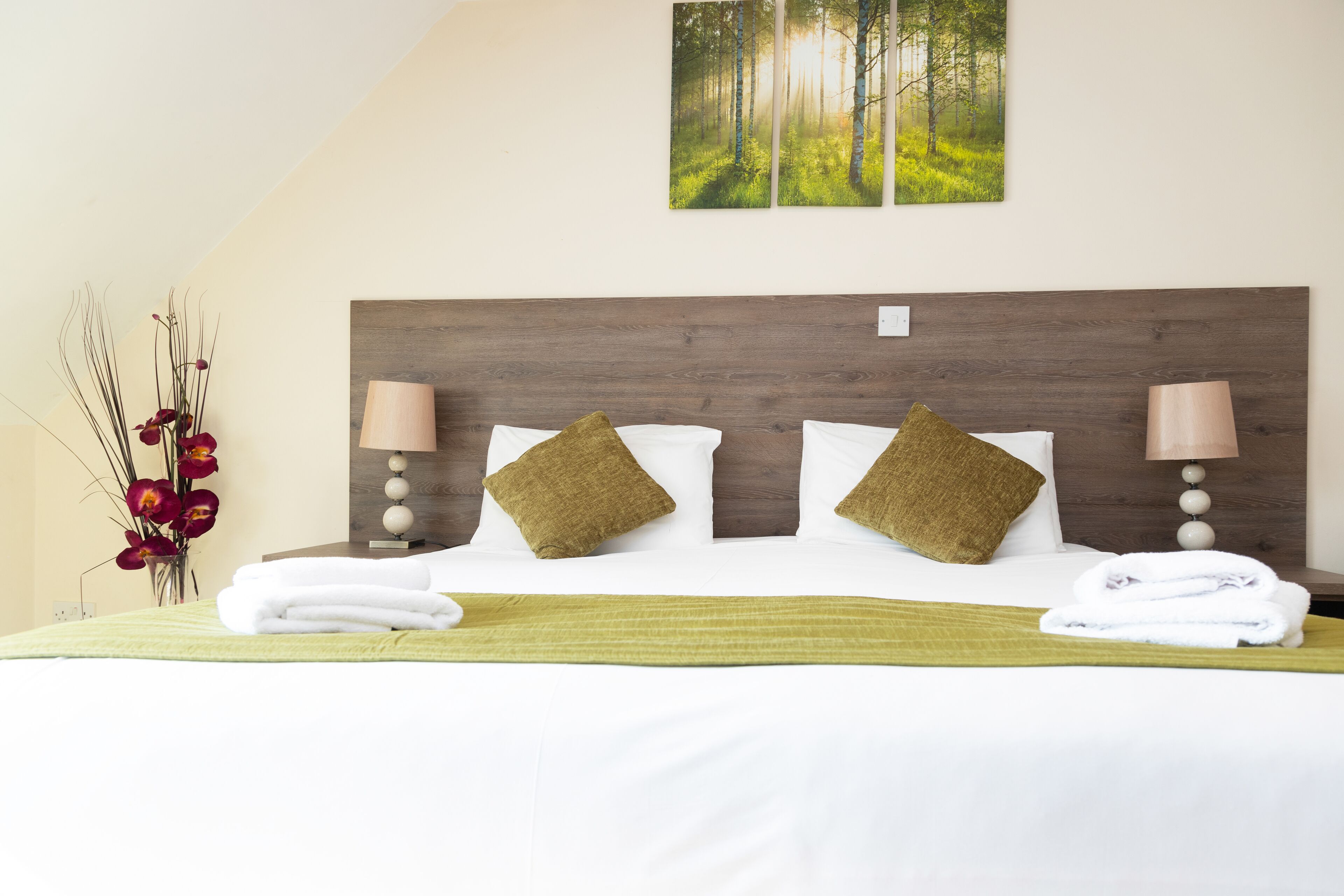A neatly made bed with white linens and two brown pillows, a bedside lamp, and a wall-mounted picture of a forest.