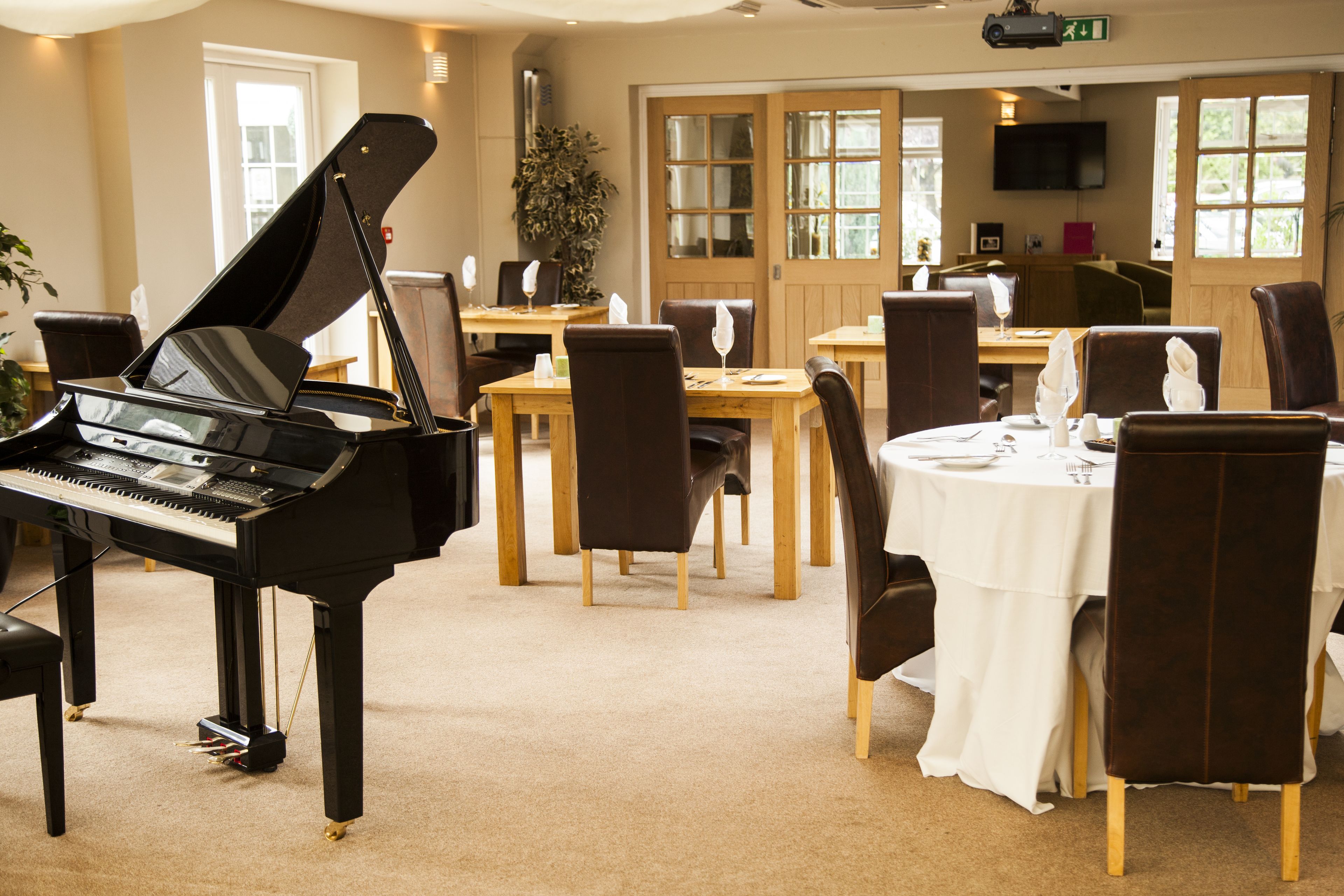 Un piano dans une pièce où se trouvent des tables et des chaises, un téléviseur accroché au mur et une porte avec des fenêtres.