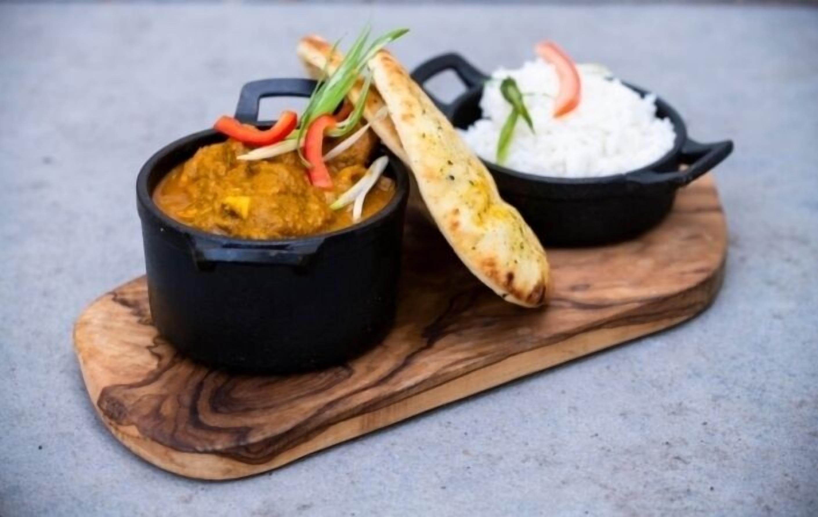 A black pot with a curry and a bowl of white rice on a wooden board.