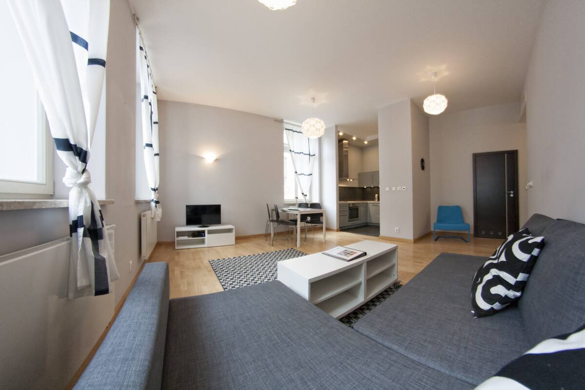 Ein modernes Wohnzimmer mit einem grauen Sofa, einem weißen Couchtisch und einem Fernseher. Es gibt einen Essbereich mit Tisch und Stühlen sowie eine Küche im Hintergrund.
