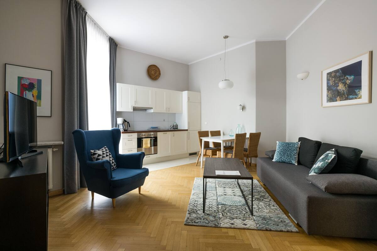 Ein modernes Wohnzimmer mit Sofa, Sessel und Esstisch. Im Hintergrund ist eine Küche zu sehen.