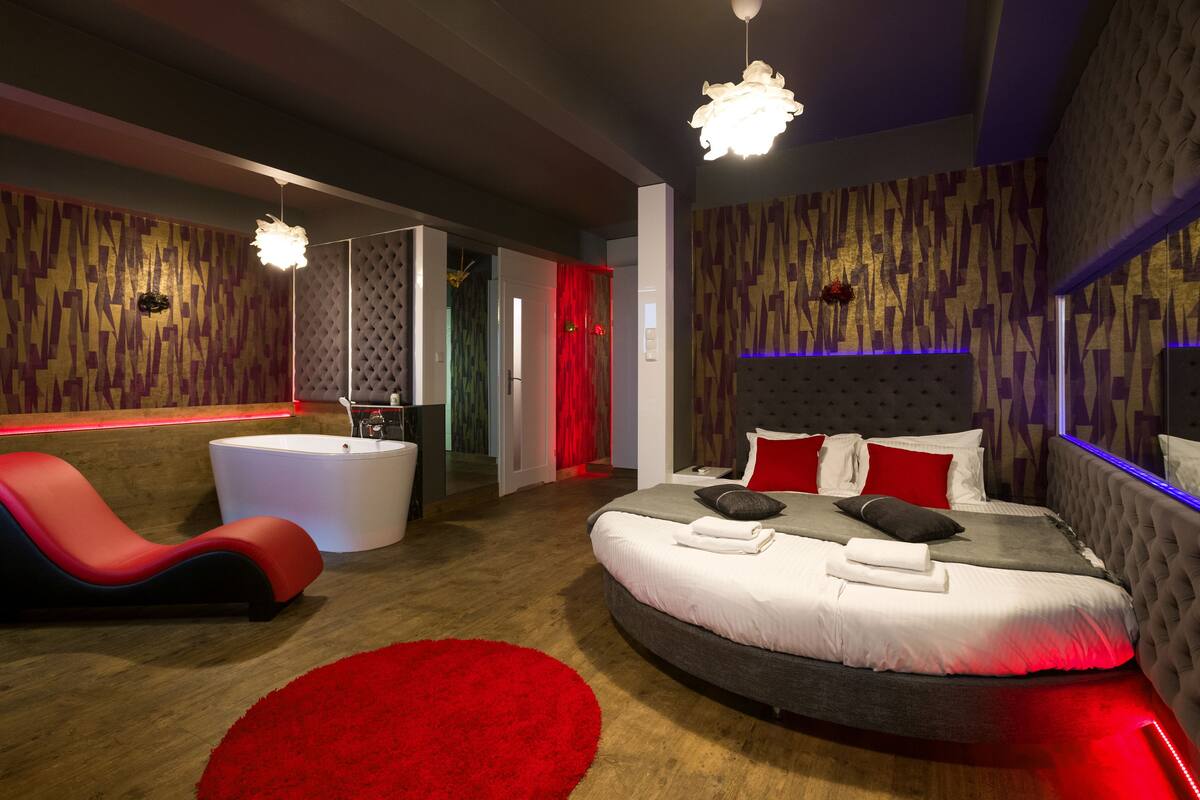 Ein modernes Schlafzimmer mit einem gewölbten Bett, einem roten Loungesessel, einer weißen Badewanne und einem roten Teppich.