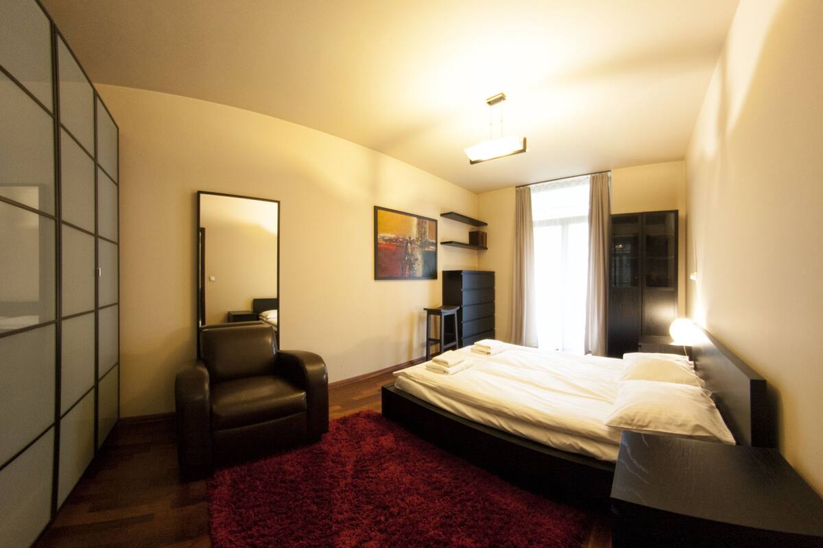 Ein Schlafzimmer mit einem Bett, einem Sessel, einer Kommode, einem Spiegel und einem roten Teppich.
