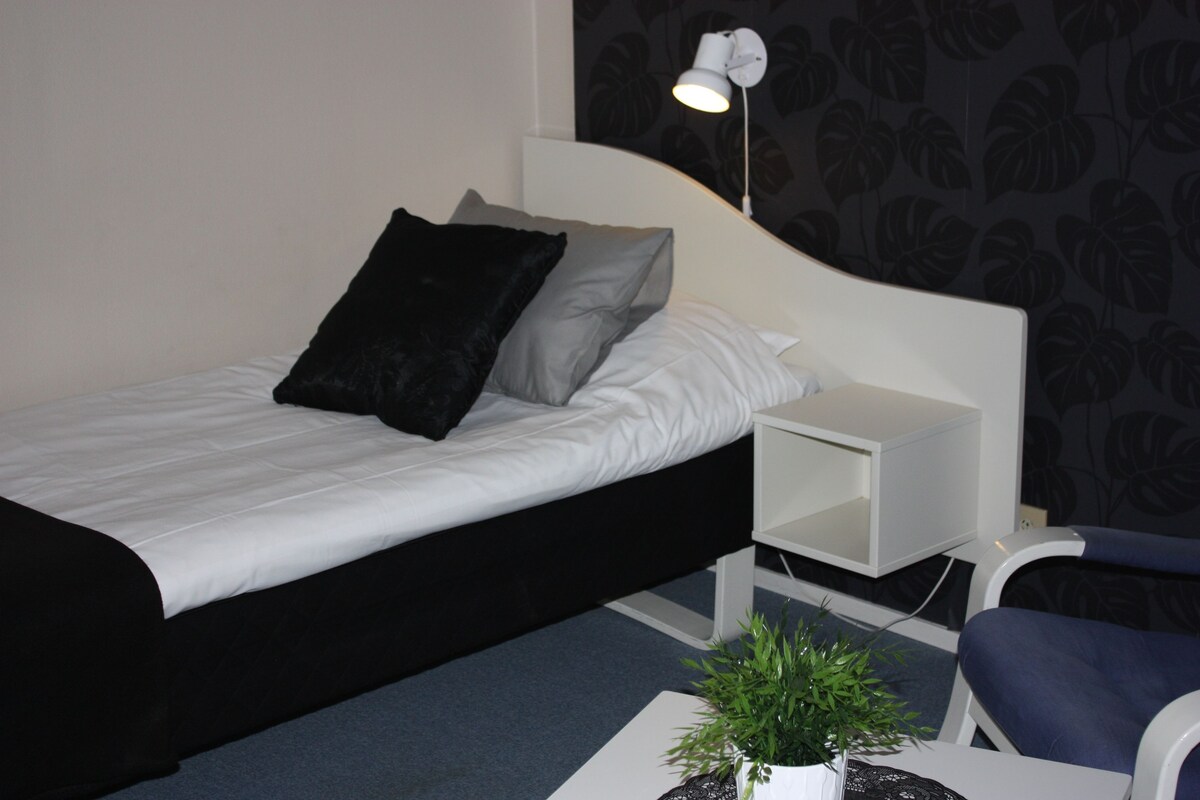 A neatly made bed with white and black bedding, a small bedside table with a lamp, and a chair with a dark cushion.