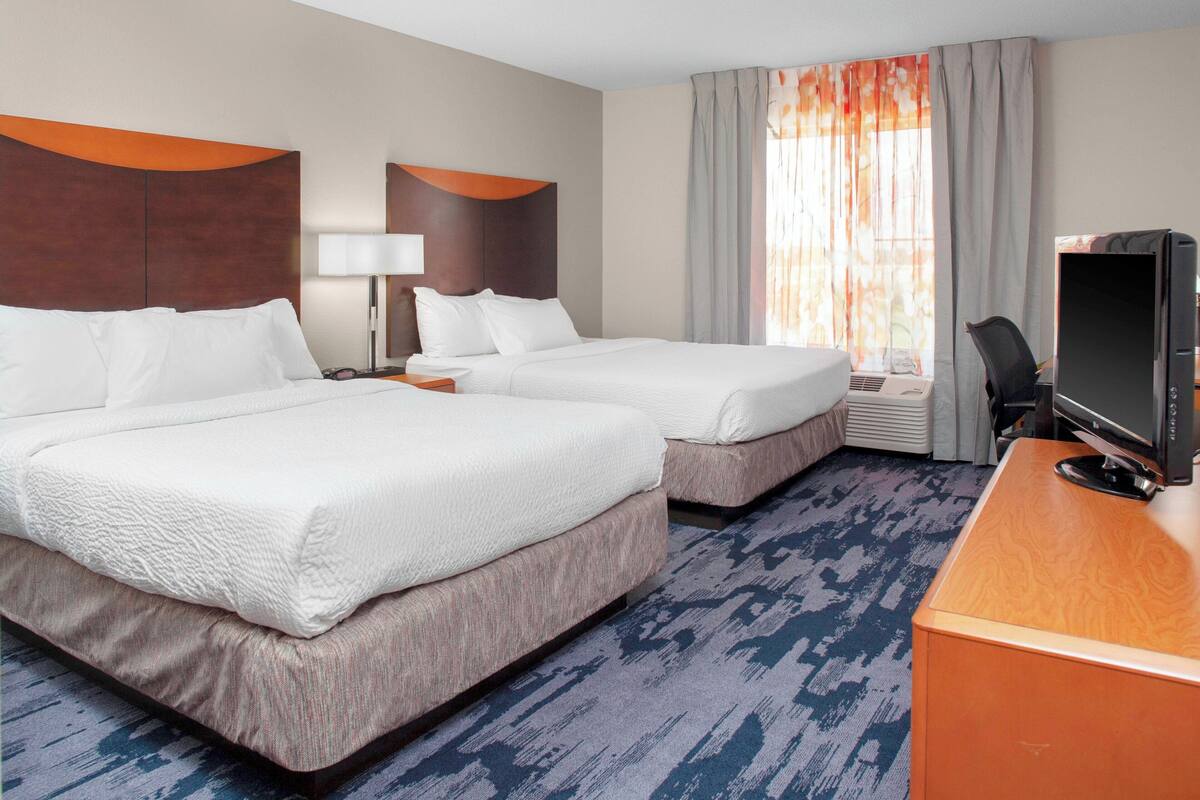 A hotel room with two beds, a desk with a computer, a lamp, and a window with curtains.