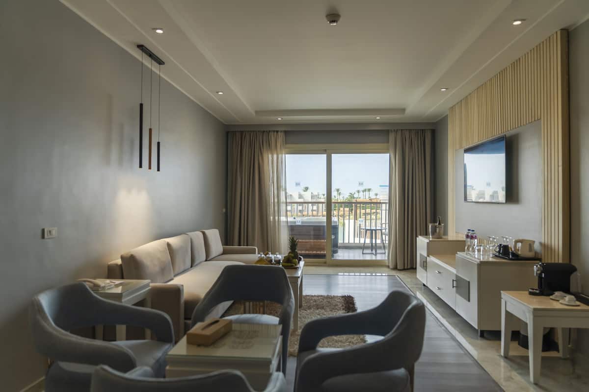 Una habitación de hotel moderna con sofá, sillas, una mesa de comedor, televisor y balcón con vistas.