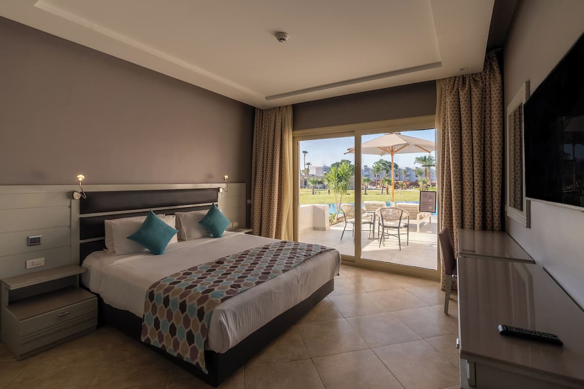 Habitación de hotel con una cama grande, un escritorio, un televisor y un ventanal corredero que da a un espacio exterior con piscina y sombrillas.