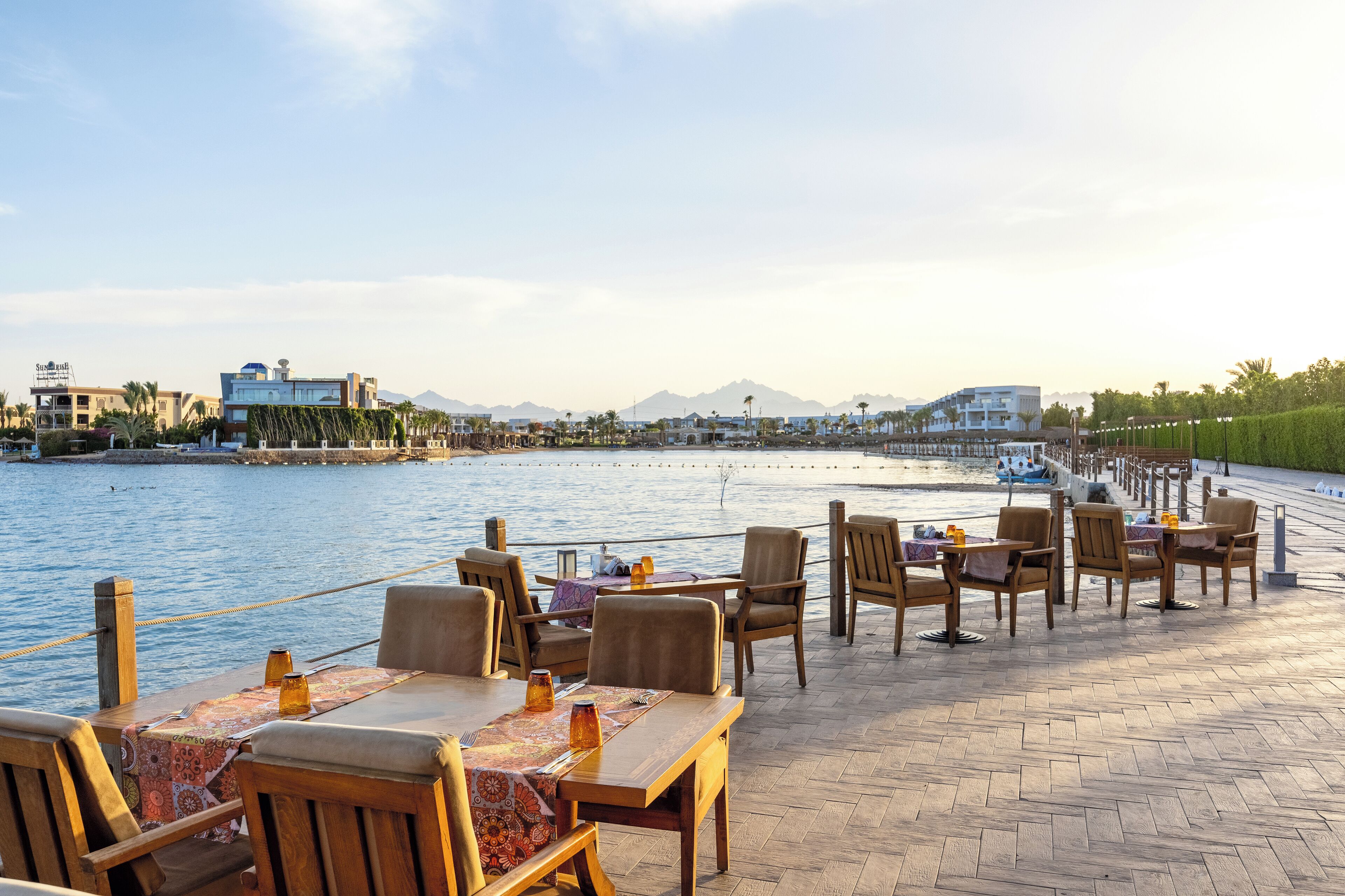 An outdoor dining area with wooden tables and chairs, overlooking a body of water with boats and buildings in the background.