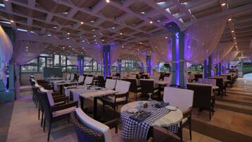 A spacious restaurant with multiple tables and chairs, a high ceiling with wooden beams, and large windows allowing natural light.