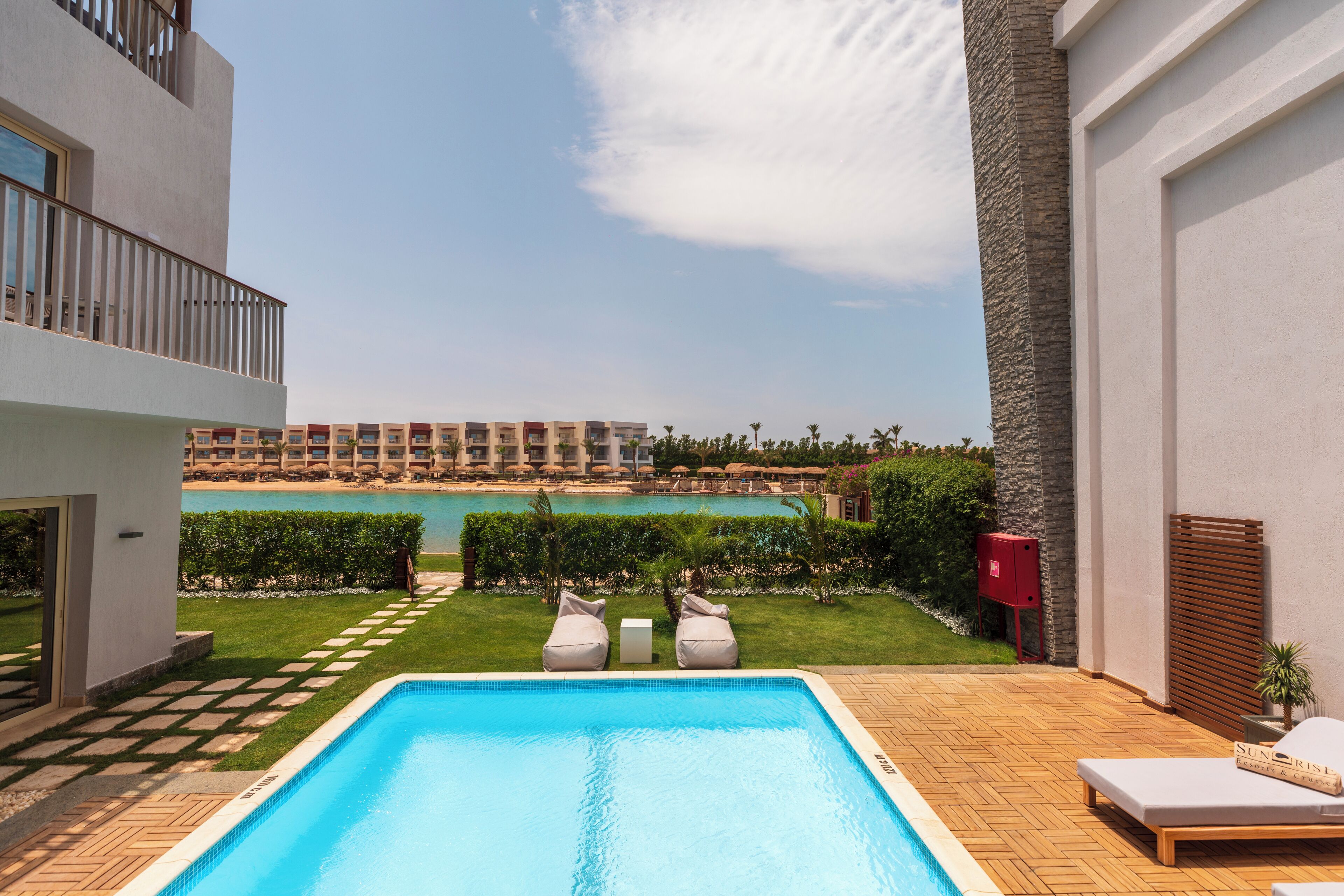 A modern outdoor pool area with lounge chairs, a wooden deck, and a view of a hotel building and beach in the background.