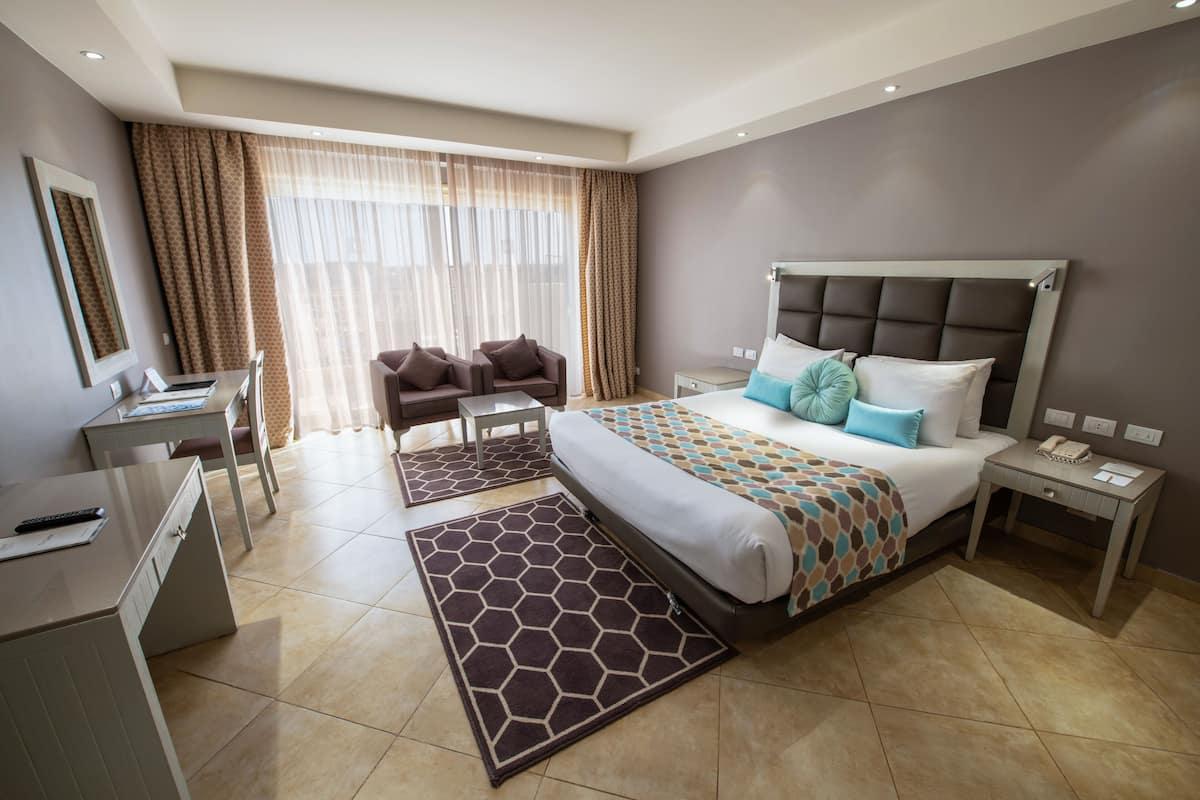 Habitación de hotel con una cama, dos sillas, un escritorio, una mesita de noche y un amplio ventanal con cortinas.