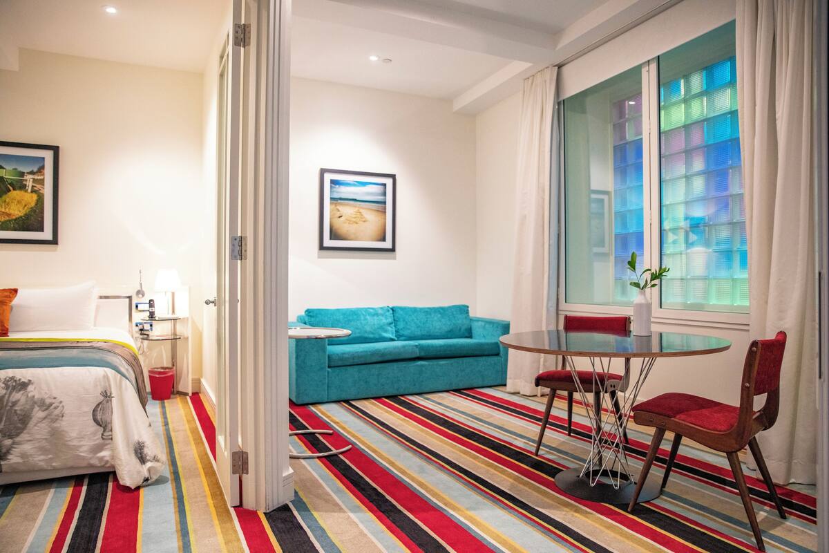 A hotel room with a bed, a blue sofa, a dining table with chairs, a striped carpet, and a large window with curtains.