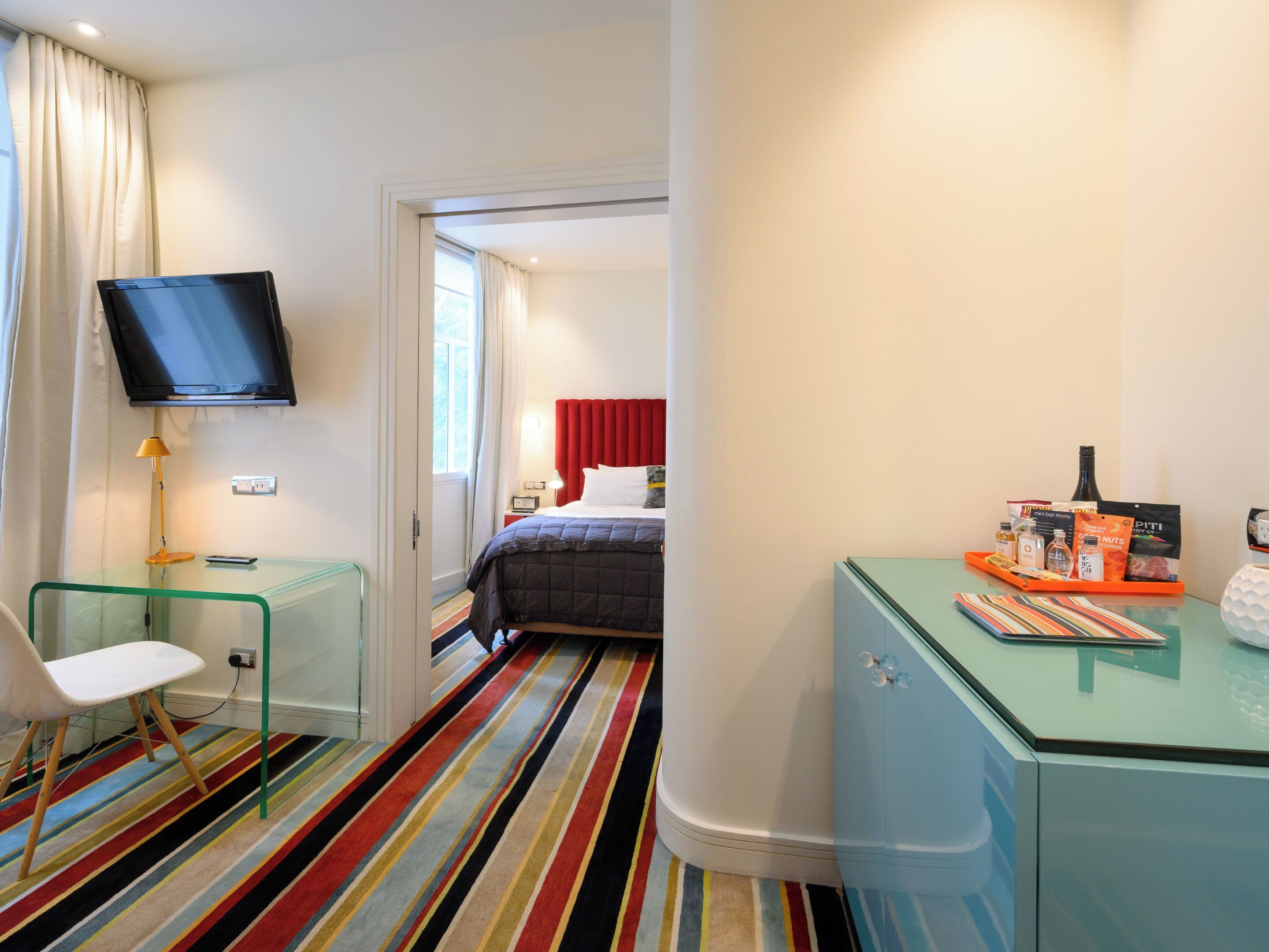 A hotel room with a bed, a TV mounted on the wall, a desk with a chair, and a colorful striped carpet.
