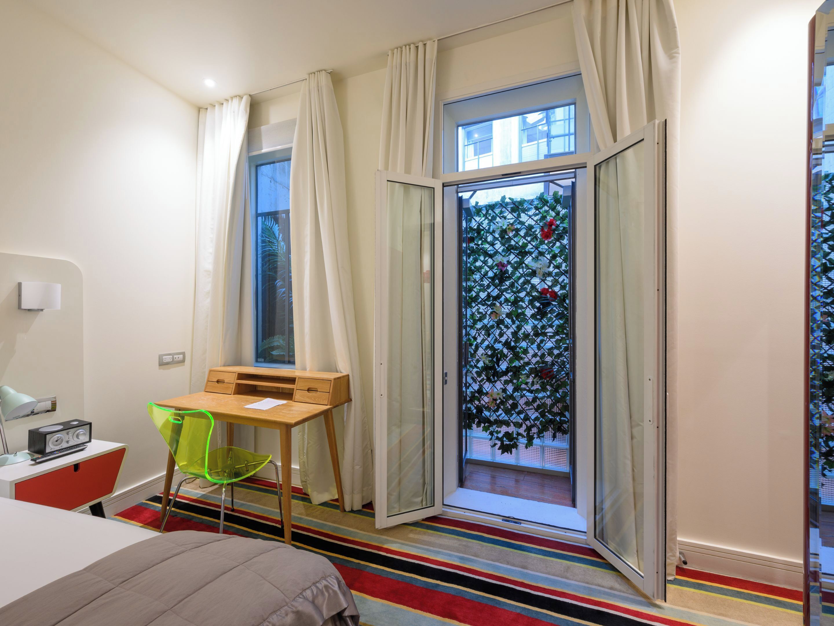 Ein modernes Hotelzimmer mit Bett, Schreibtisch mit Stuhl, einem Fenster mit Vorhängen und Blick ins Grüne.