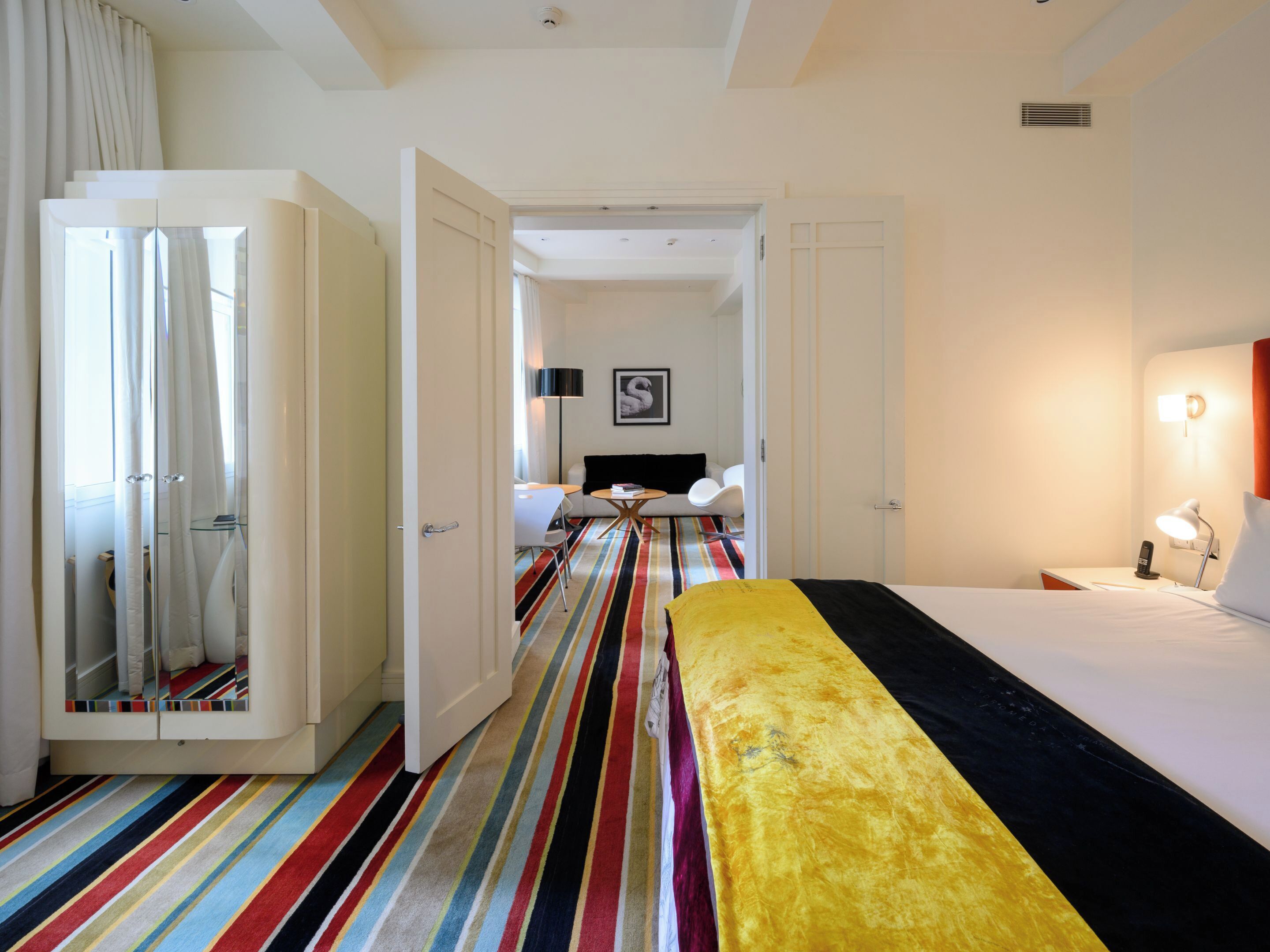 A hotel room with a bed, a desk, a chair, a lamp, and a view of the hallway.