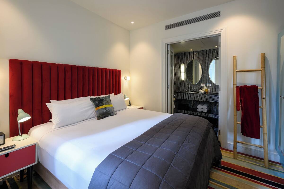 A hotel room with a large bed, a red headboard, a bedside table with a lamp, and a bathroom visible through an open door.
