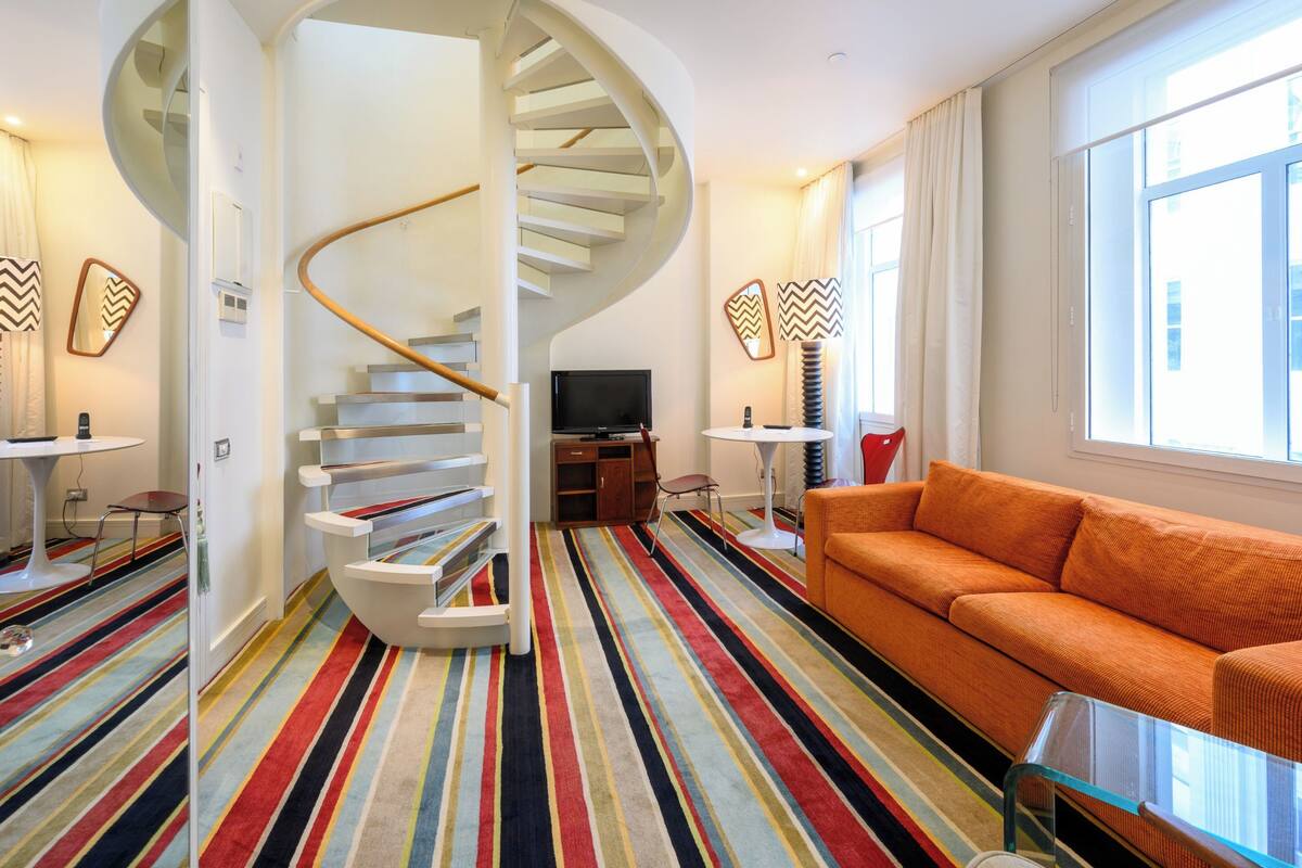 A modern living room with a spiral staircase, a colorful striped carpet, an orange sofa, and a TV.