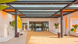 Een moderne hotelentree met een glazen luifel, potplanten en een vlag.