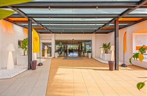 A modern hotel entrance with a glass canopy, potted plants, and a flag. - Hotel Caribe Ibiza (Santa Eulalia del Rio)