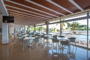 A covered outdoor seating area with tables and chairs, large windows, and a pool view. - Hotel Caribe Ibiza (Santa Eulalia del Rio)