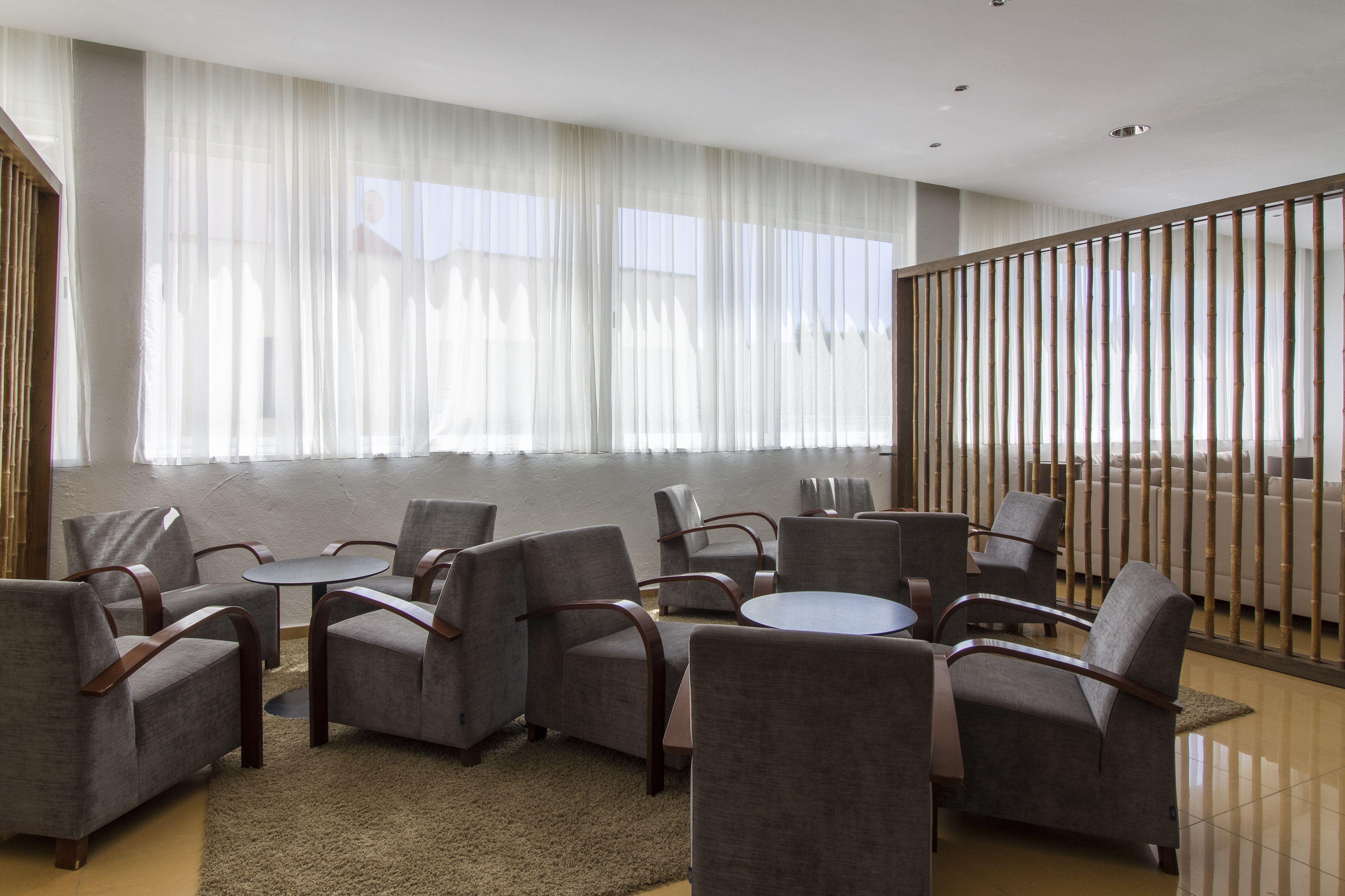A modern hotel room with a seating area featuring grey upholstered chairs and a round table, large windows with sheer curtains, and a view of the beach.