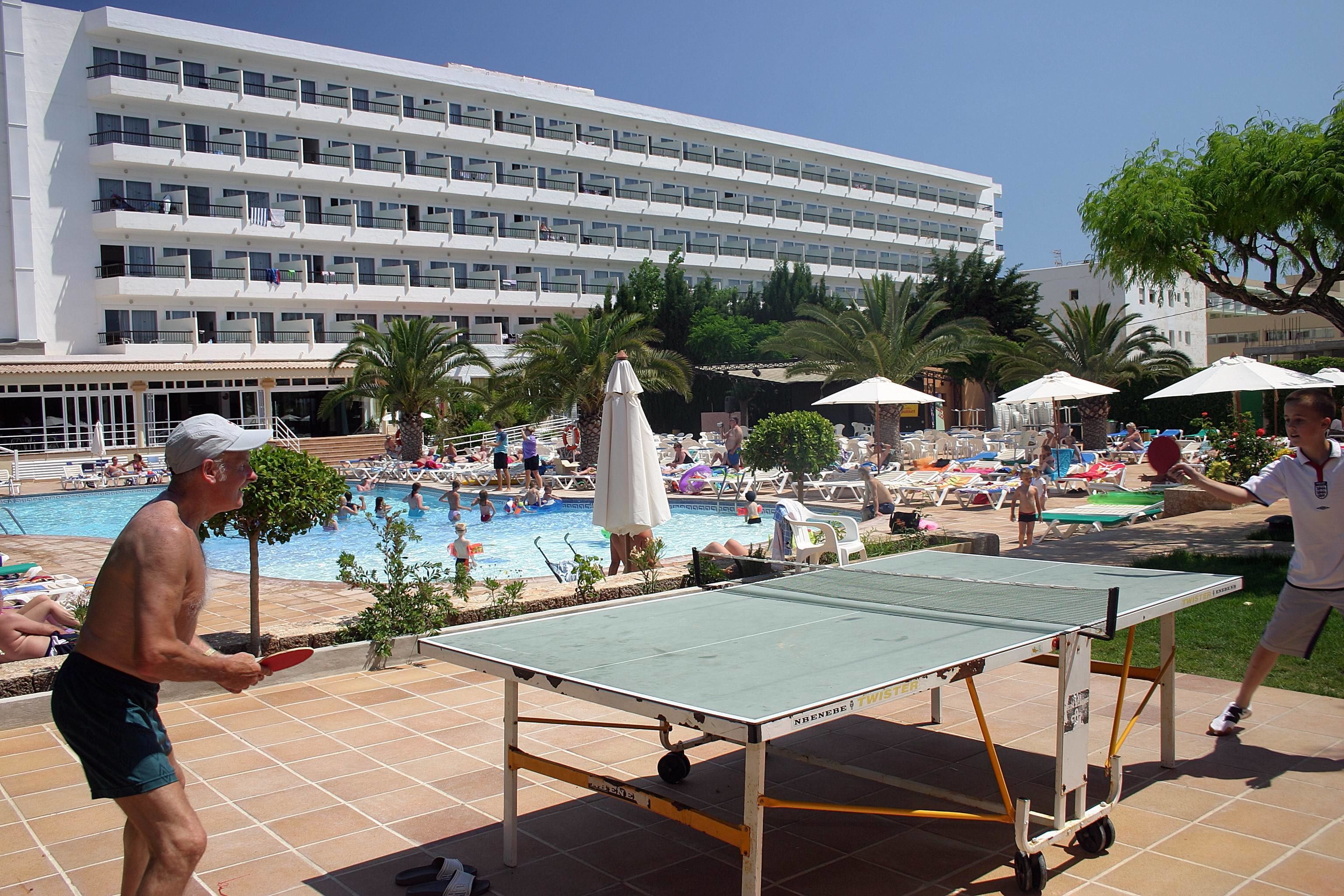 A pool area with a ping pong table, lounge chairs, umbrellas, and a large building in the background.