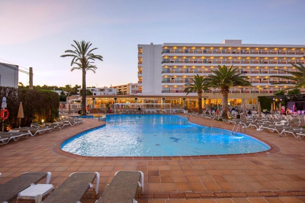 Ein Swimmingpool mit Liegestühlen und Palmen, umgeben von einem Hotelgebäude mit mehreren Balkonen.