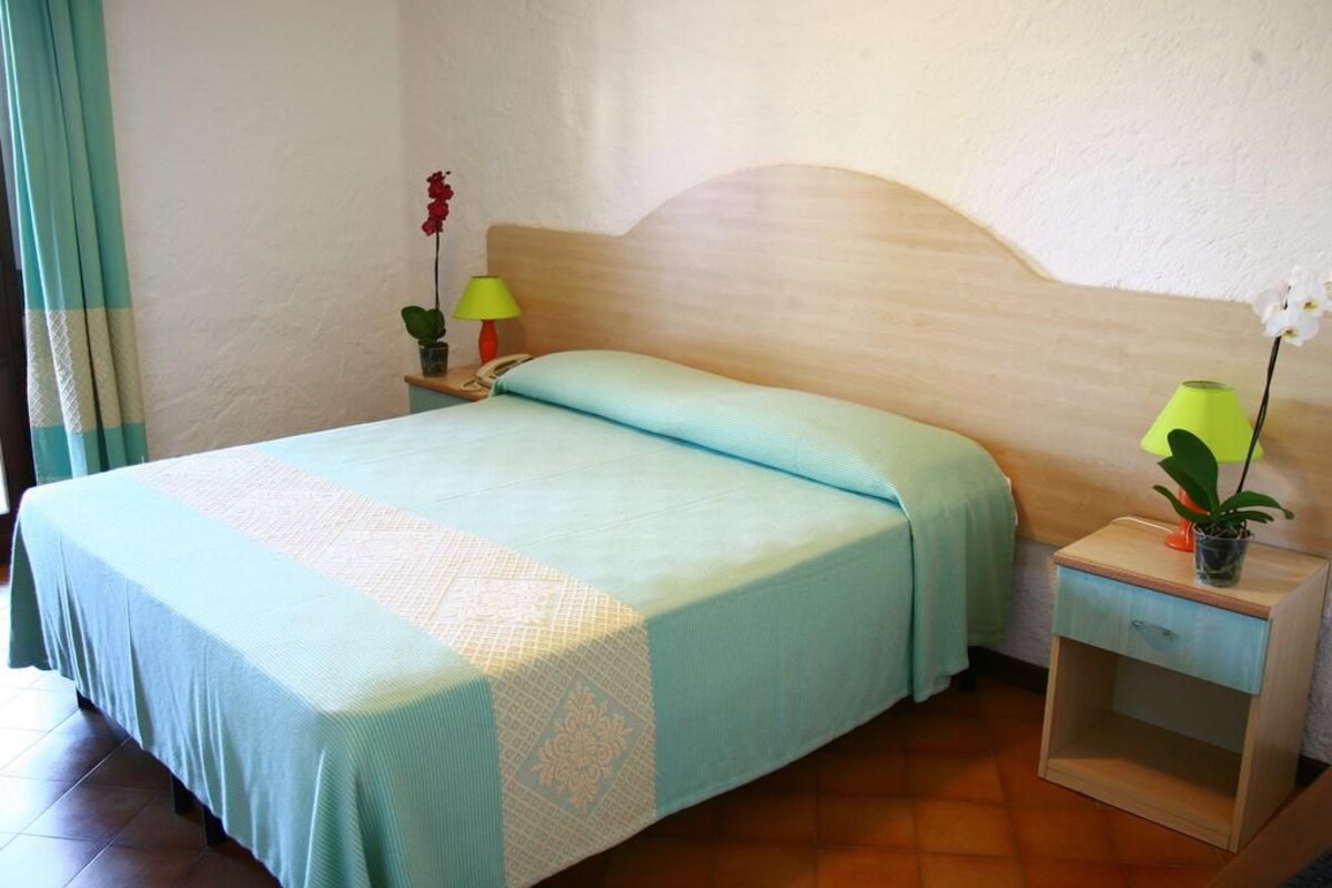 A single bed with a light blue bedspread and a wooden headboard. There is a nightstand with a lamp and a small potted plant.