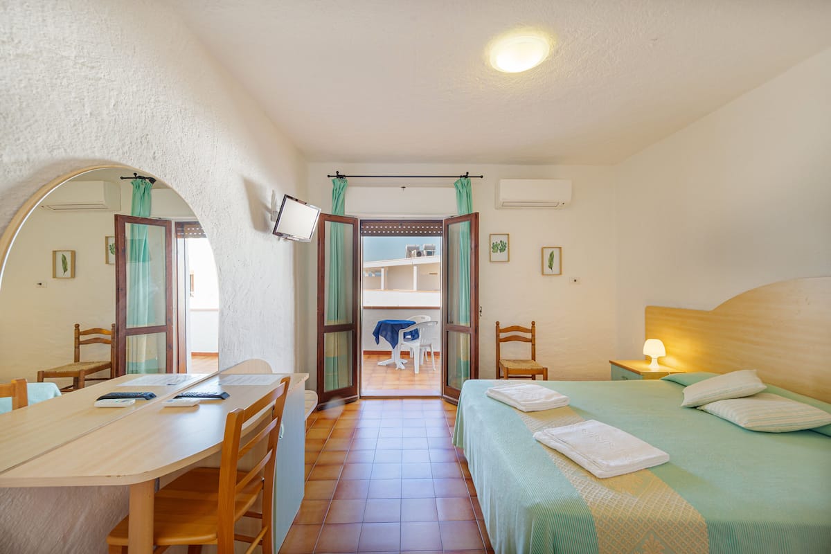 Ein Zimmer mit einem Bett, einem Schreibtisch, Stühlen, einem Fernseher und Blick durch eine offene Tür.