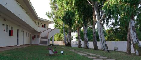 A courtyard with a staircase leading to a covered area, surrounded by trees and a white building with multiple doors.