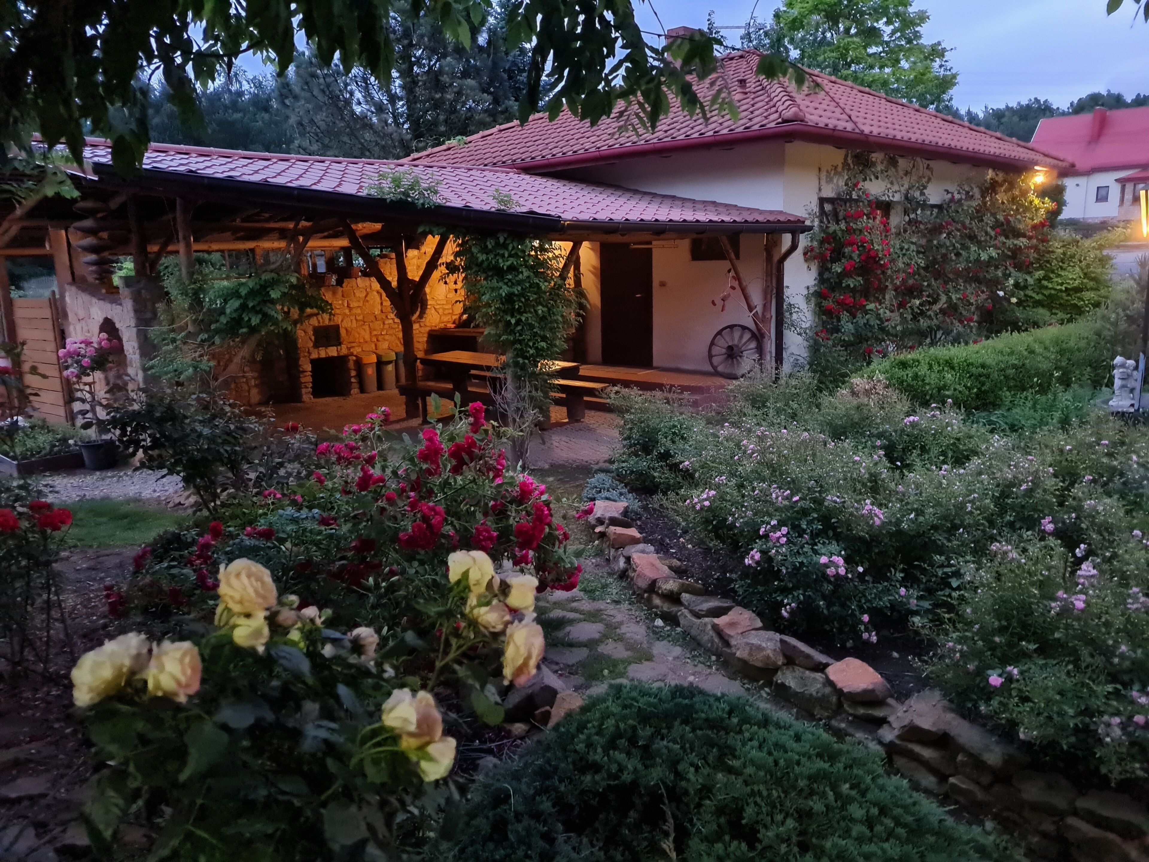 Una casa rústica con techo de tejas, pared de piedra y jardín con diversas plantas y flores.