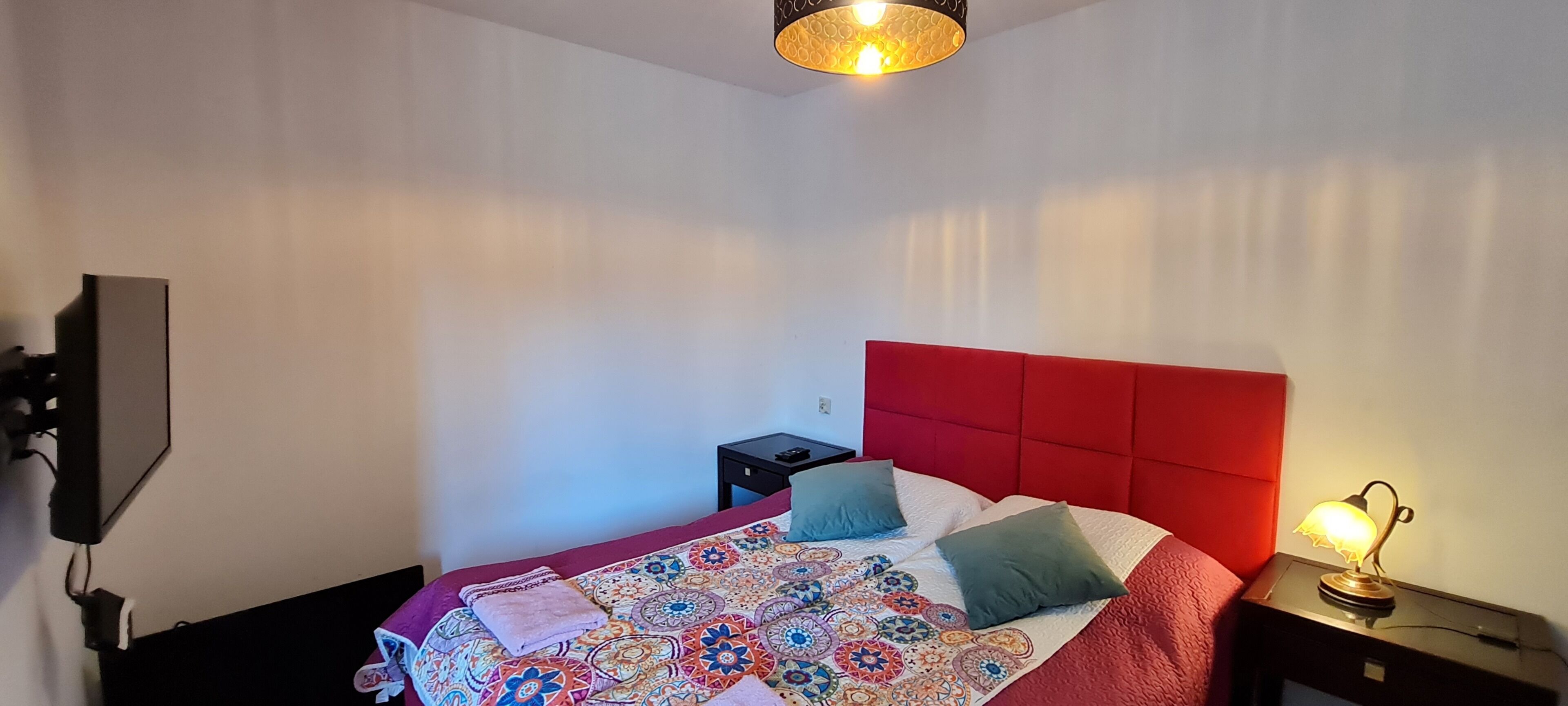 A bedroom with a red headboard, a patterned bedspread, two pillows, a bedside lamp, a small bedside table, and a wall-mounted TV.