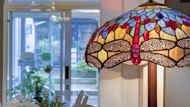 A stained glass lamp with a geometric pattern, a white chair, and a view of greenery outside a window. - Avoca House (Dublin)