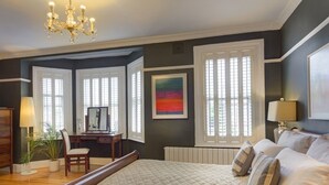 A bedroom with a large bed, a desk, and a chair. There are two windows with blinds, a chandelier, and a painting on the wall. - Avoca House (Dublin)