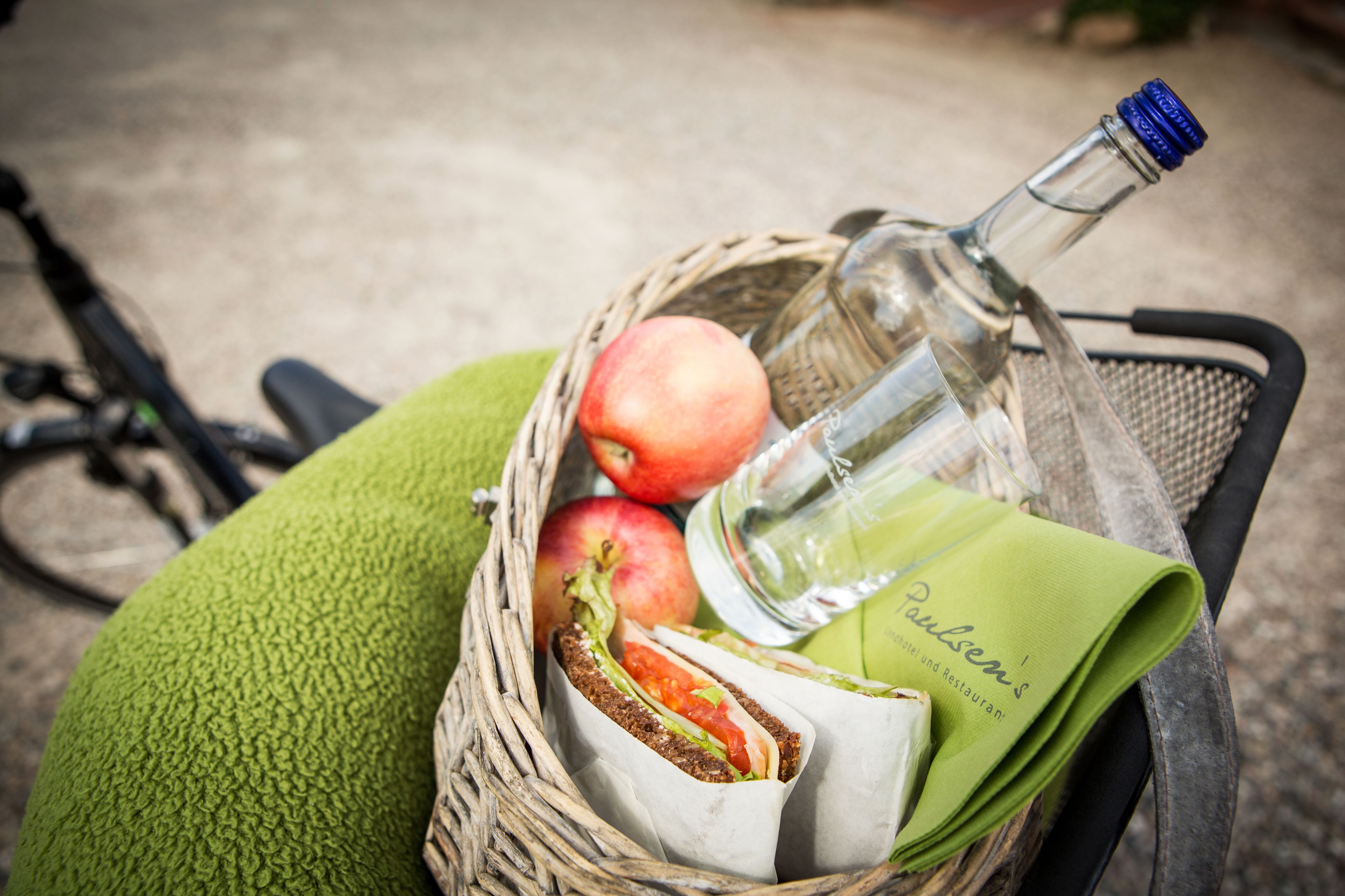 A wicker basket containing a sandwich, a bottle, and apples.