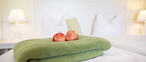 Una cama bien hecha con una colcha verde y dos manzanas encima, frente a un cabecero y mesitas de noche con lámparas.