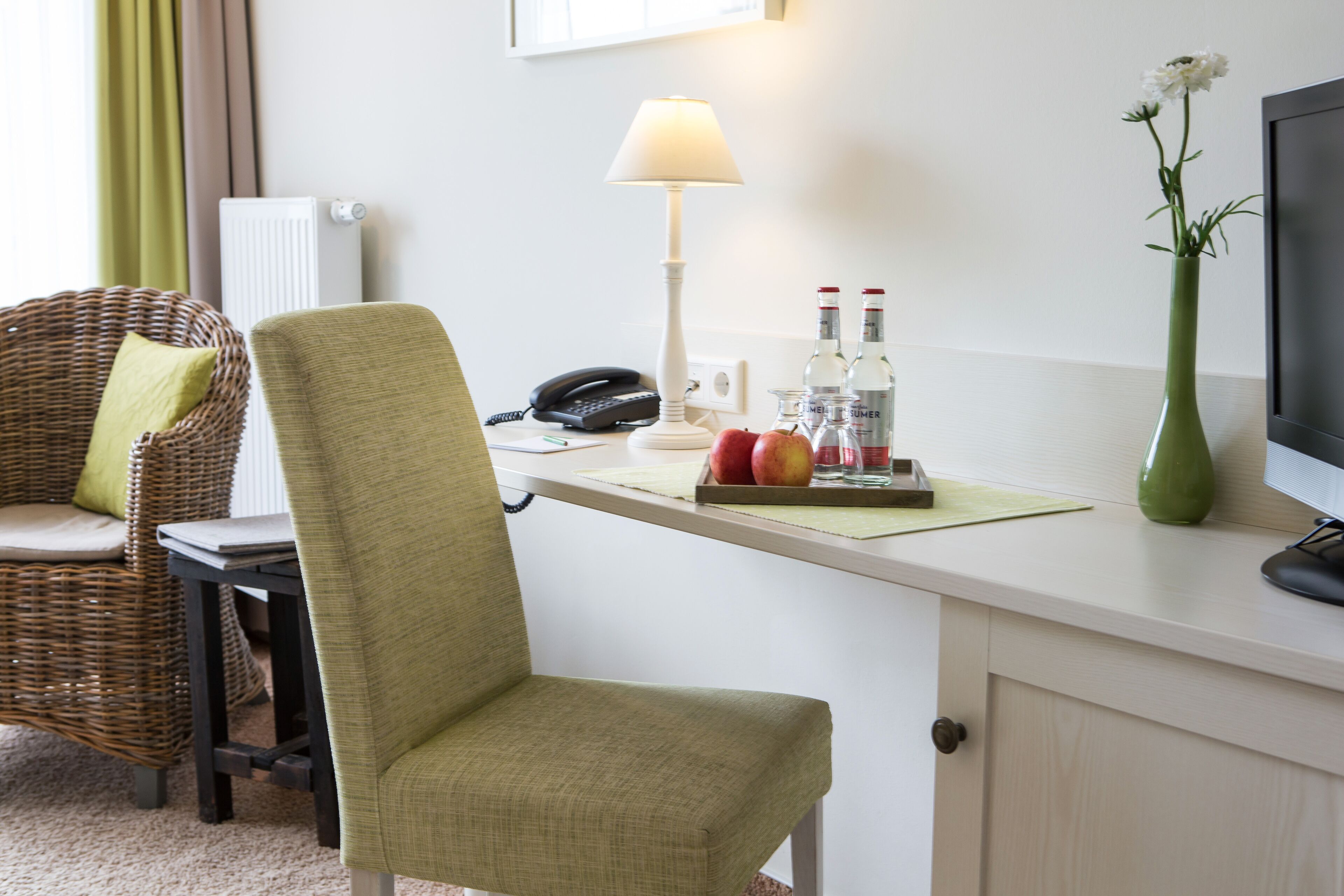 Une pièce comprenant un fauteuil en osier, un bureau avec une lampe, un vase de fleurs et un téléviseur.