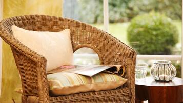 Une chaise en osier, dotée d’un coussin et accompagnée d’un magazine, est placée près d’une fenêtre offrant une vue sur la verdure.